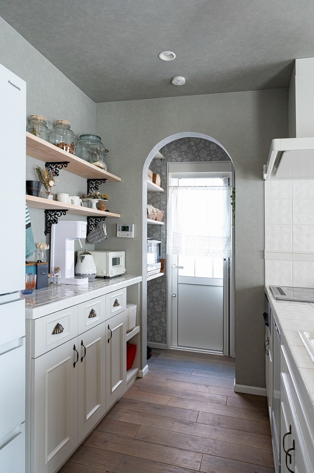 キッチンはタイルの貼り方や水栓やシンク・食洗器などの機器にもこだわり造作しました。

框を取り入れたデザインの扉にすることでよりフレンチシャビーなインテリアを演出します。アーチ下がり壁の奥のパントリーには勝手口を設け、両脇には掃除機やレンジ・食材などをたっぷり置ける可動棚があり、アクセントクロスで仕上げました。カップボードもホームランドのオリジナルで、施主支給されたブラケットを使った棚には可愛いキッチン雑貨を飾って楽しめます。可愛いだけでなく、炊飯器を引き出して使える機能性も重視しました。